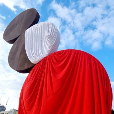 Large abstract sculpture draped in red and white fabric under a partly cloudy blue sky.