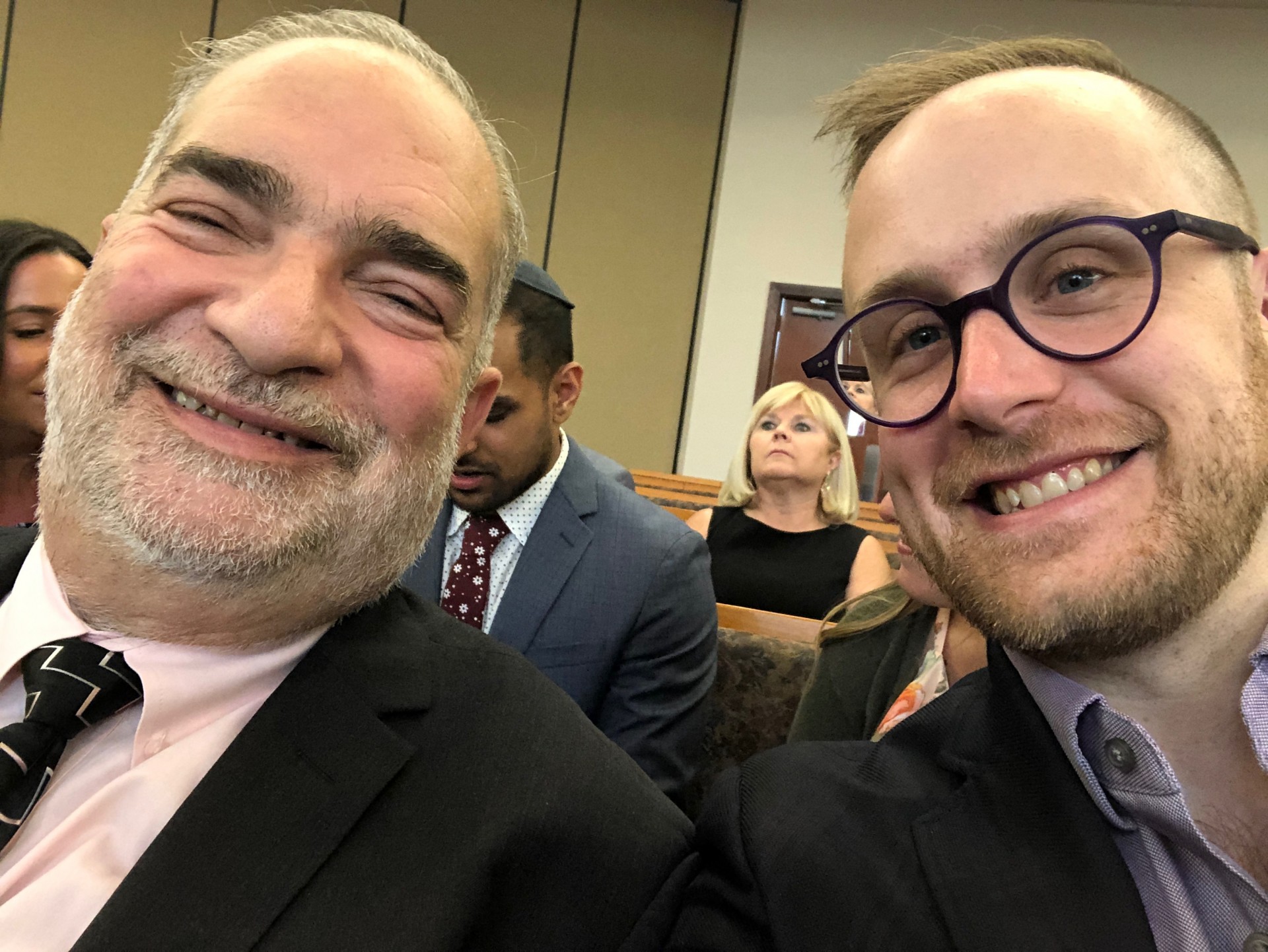 Two smiling individuals in suits take a selfie at an indoor event, with people seated in the background.