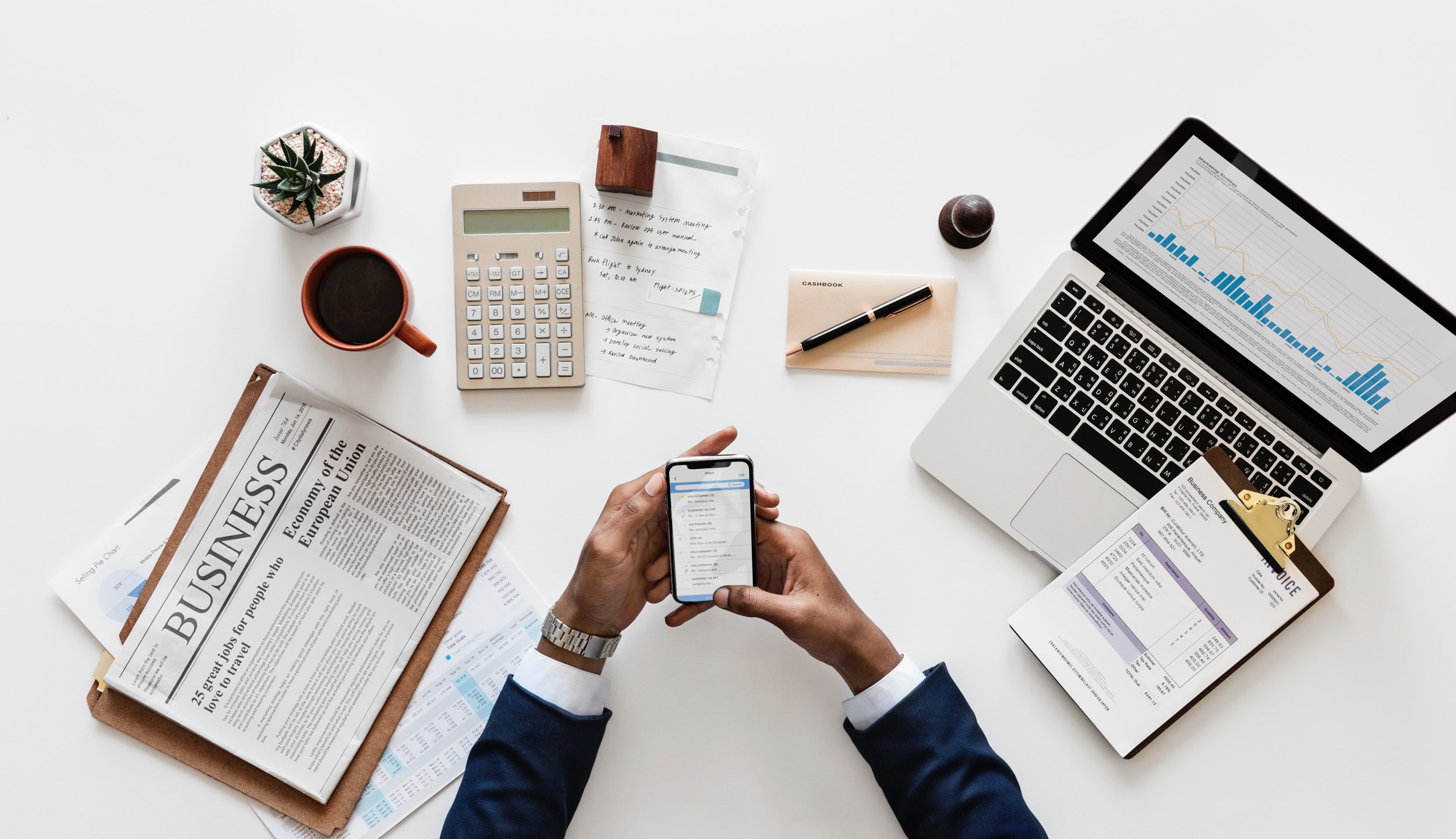 arms of person in a suit, using smartphone, surrounded with laptop, calculator, coffee cup, newspaper