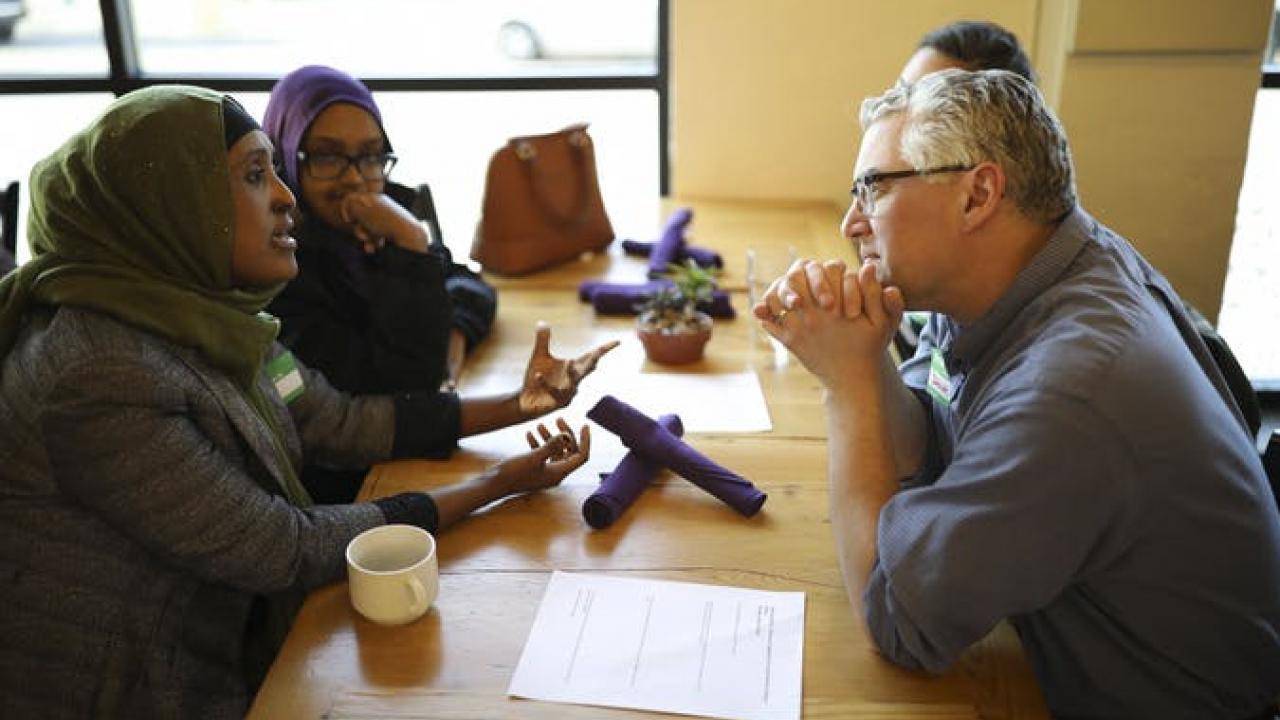 A diverse group of people sitting at a table in a discussion, with papers and a coffee cup in view.