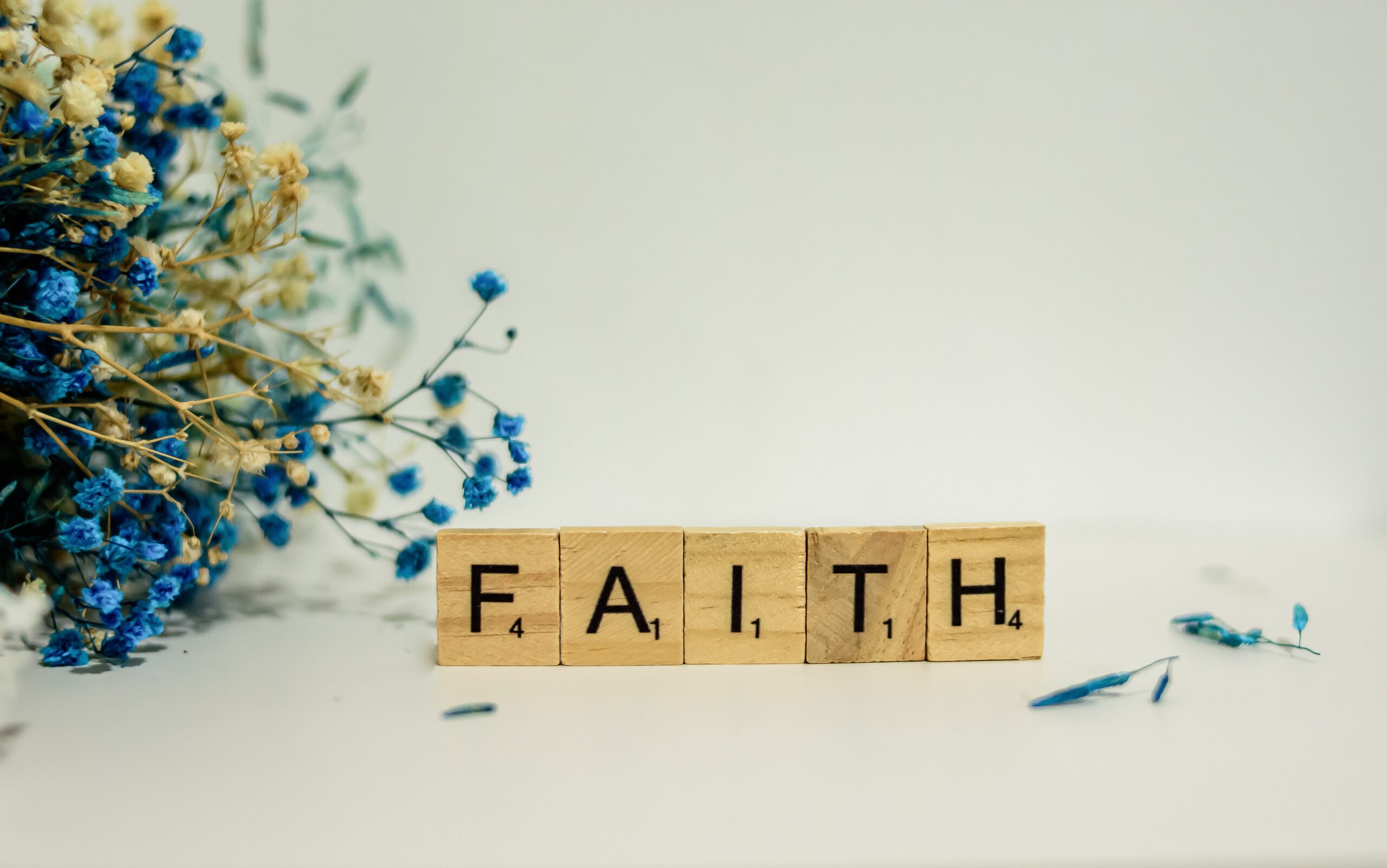 Scrabble tiles spell FAITH beside a bunch of dried blue and beige flowers on a white background.