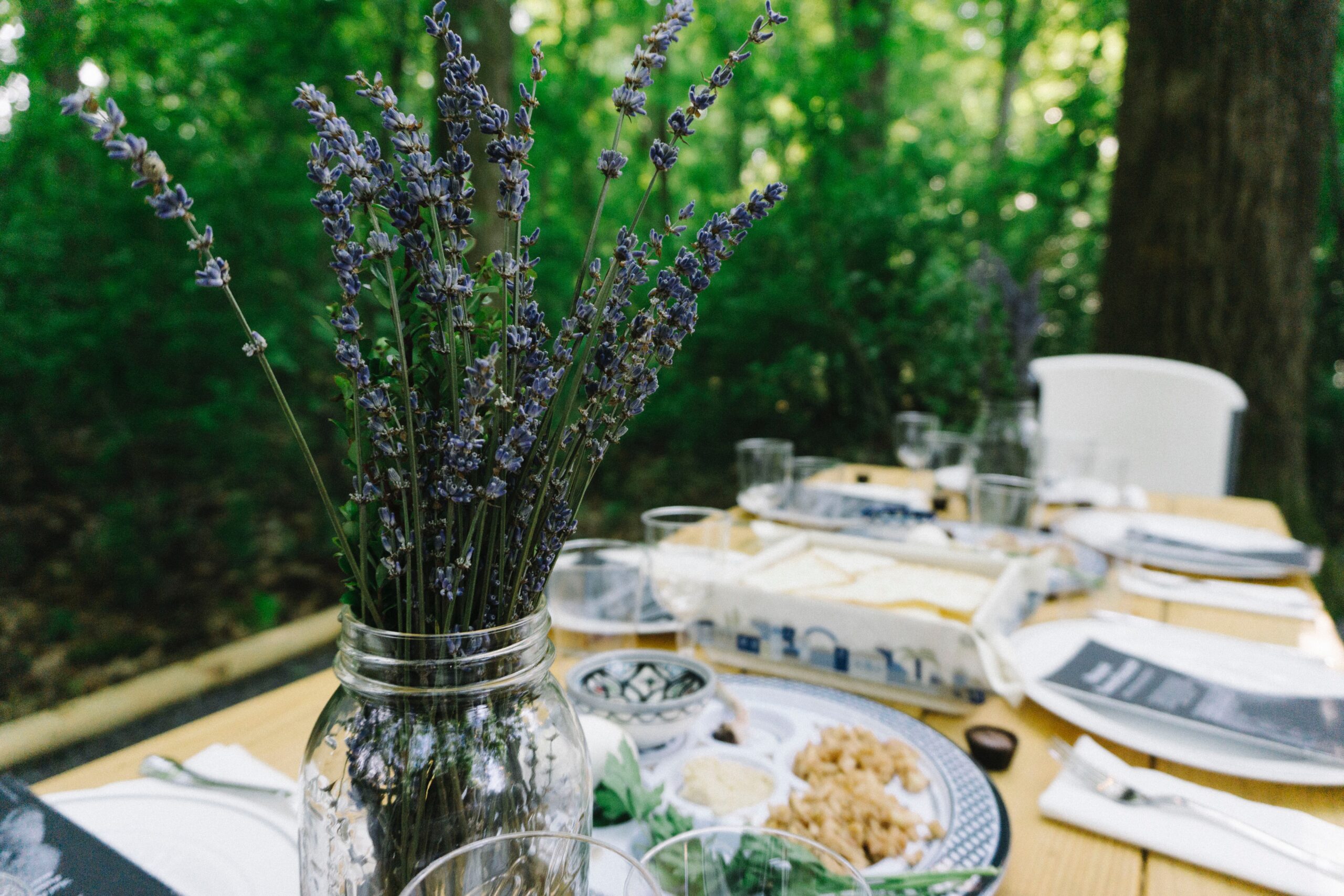 A rustic outdoor table setting with lavender in a jar and plates of food surrounded by lush green foliage.