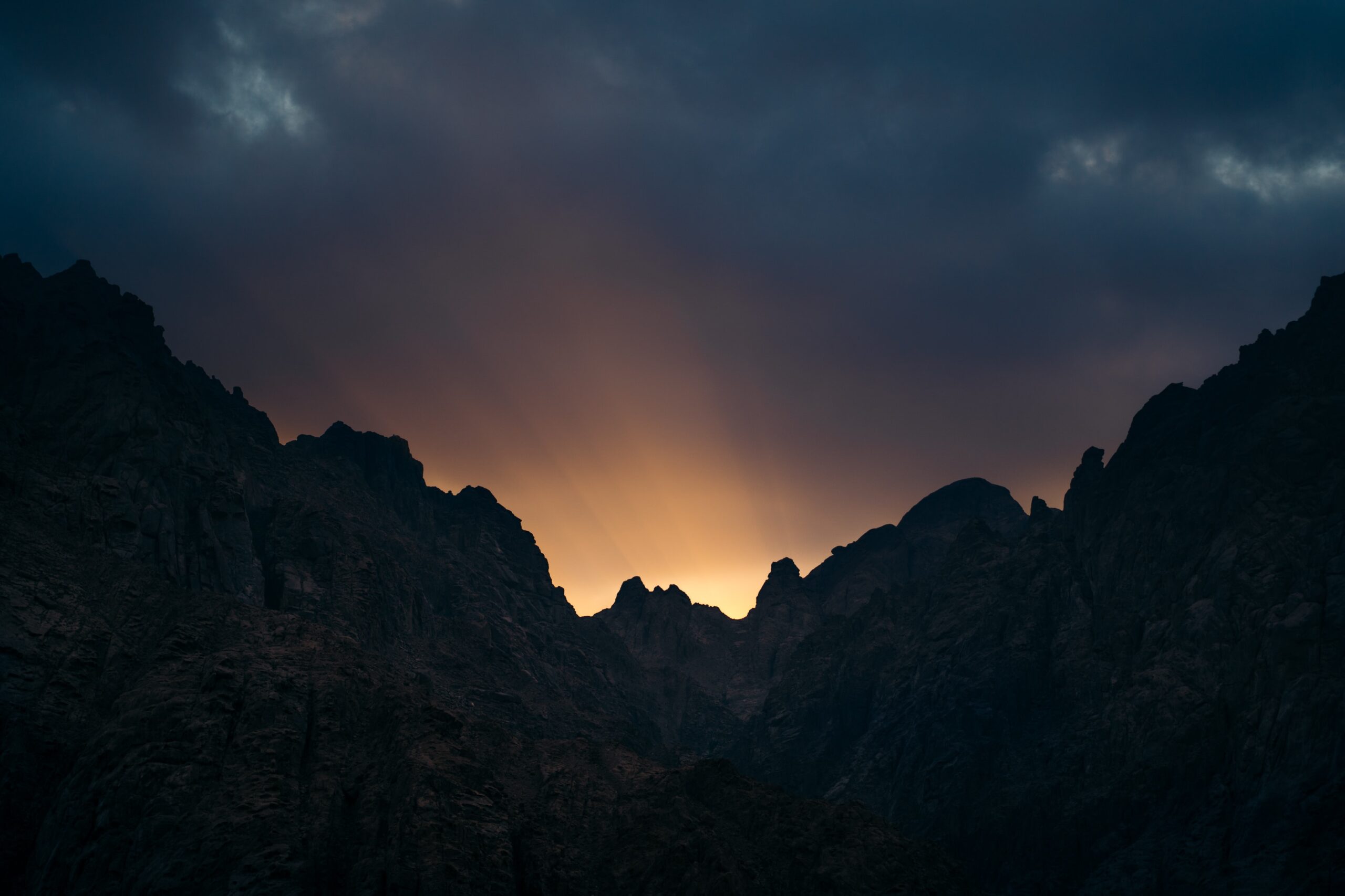 Sunset over silhouetted mountains with dramatic clouds and rays of light streaming through.