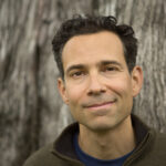 A man with short curly hair smiles slightly while standing in front of a tree trunk.