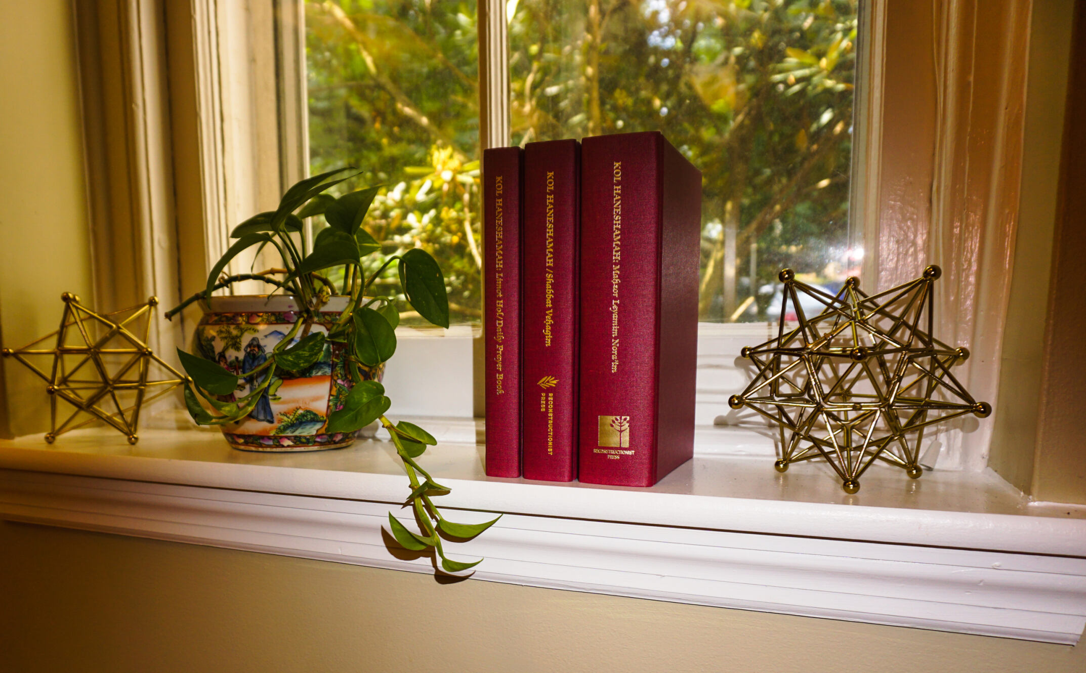 Books, a potted plant, and geometric sculptures on a window sill with a garden view.