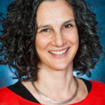 A smiling person with curly hair wearing a red top and a necklace against a blue background.