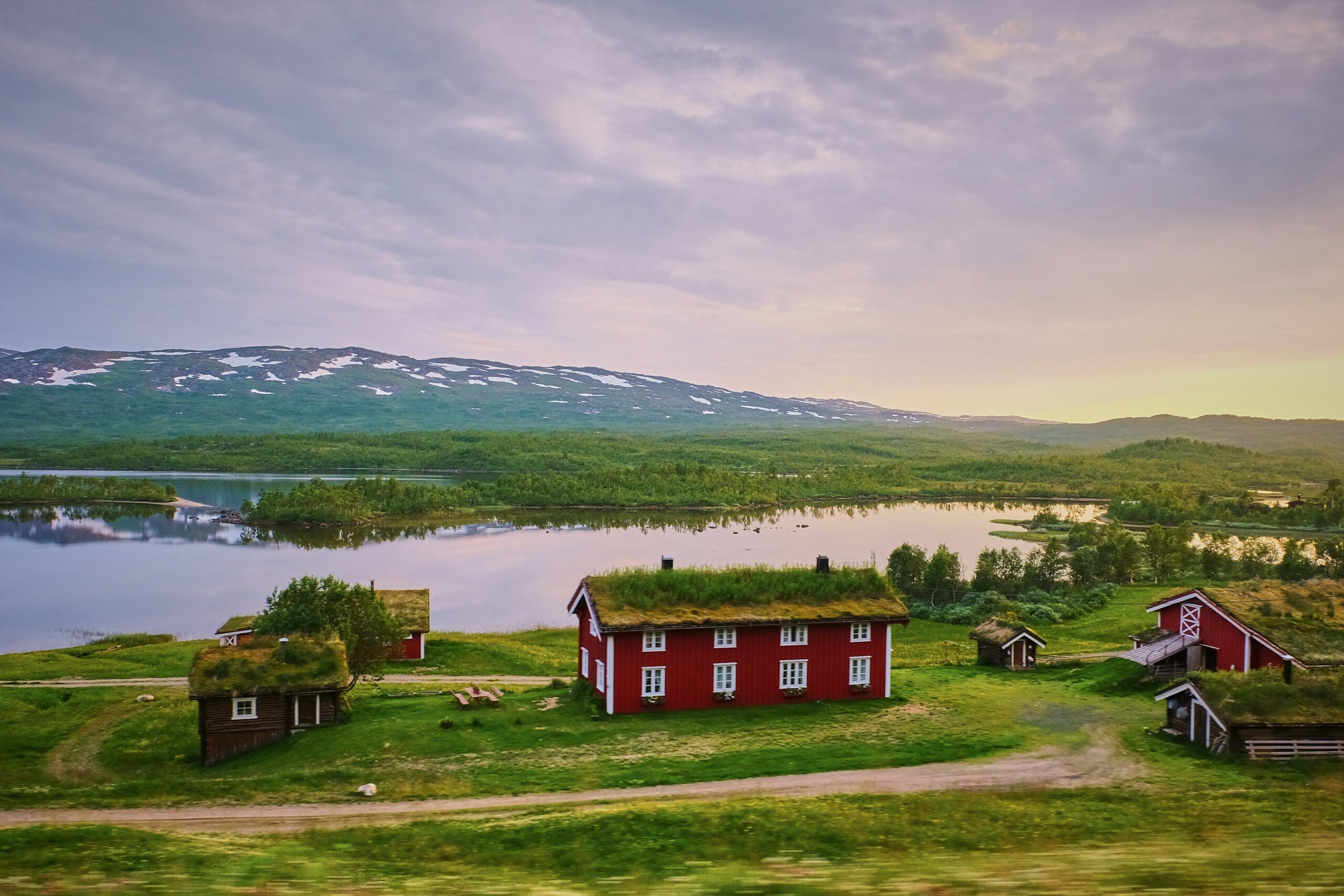 Red houses with grassy roofs beside a lake and mountains under a cloudy sky.