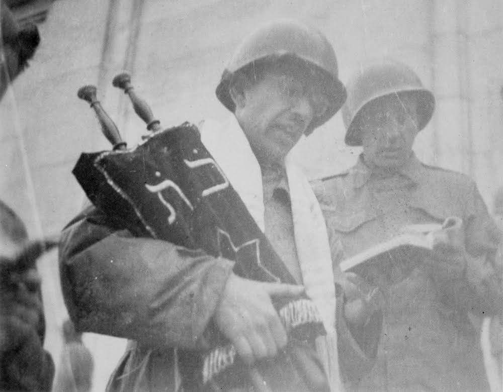 A soldier in a helmet holding a Torah scroll, accompanied by another soldier in uniform, both are reading.