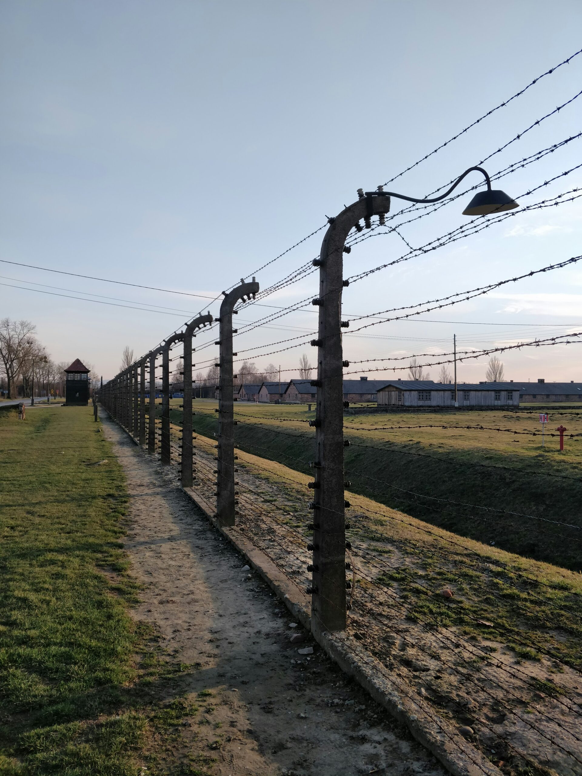 A barbed wire fence with a lamp, pathway, and guard tower along a grassy area under a clear sky.