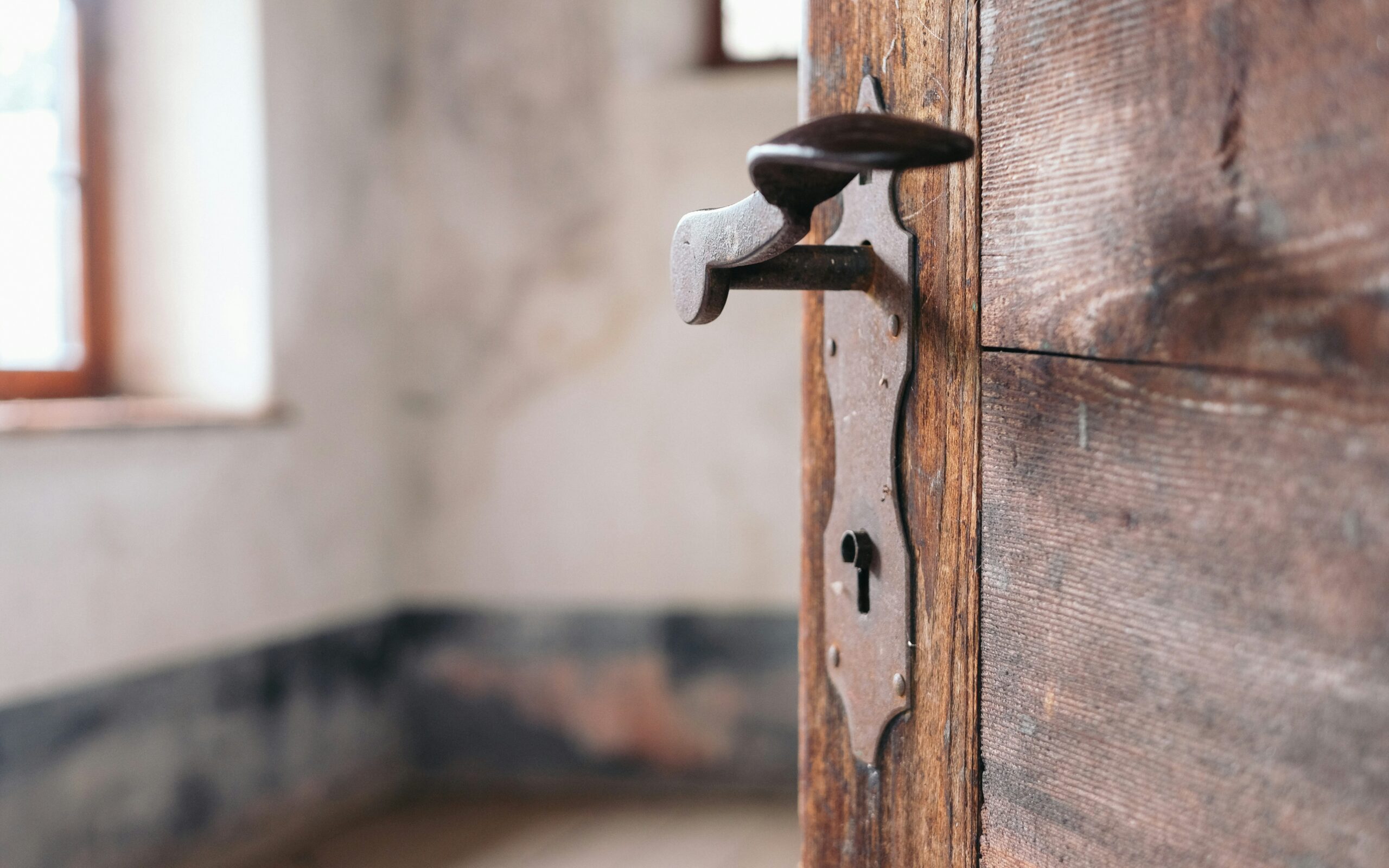 Close-up of an old wooden door with a vintage handle and keyhole, leading into an empty, blurred room.