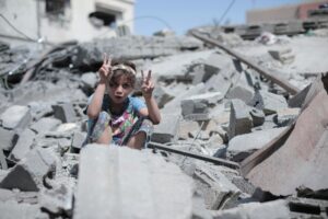 A child making peace signs sits amidst rubble and debris under a clear blue sky.