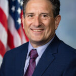 Man in a blue suit and purple tie smiling, with a U.S. flag in the background.