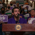 Person speaking at a podium in Pennsylvania, surrounded by people holding Ban Bribes signs.