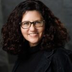 A person with curly hair and glasses smiles against a dark background.