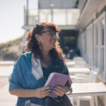 A woman with glasses smiles, holding a notebook outdoors near a building with large windows.