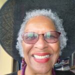 Elderly woman with curly gray hair, glasses, and a wide-brimmed hat, smiling. She wears red lipstick and beaded jewelry.