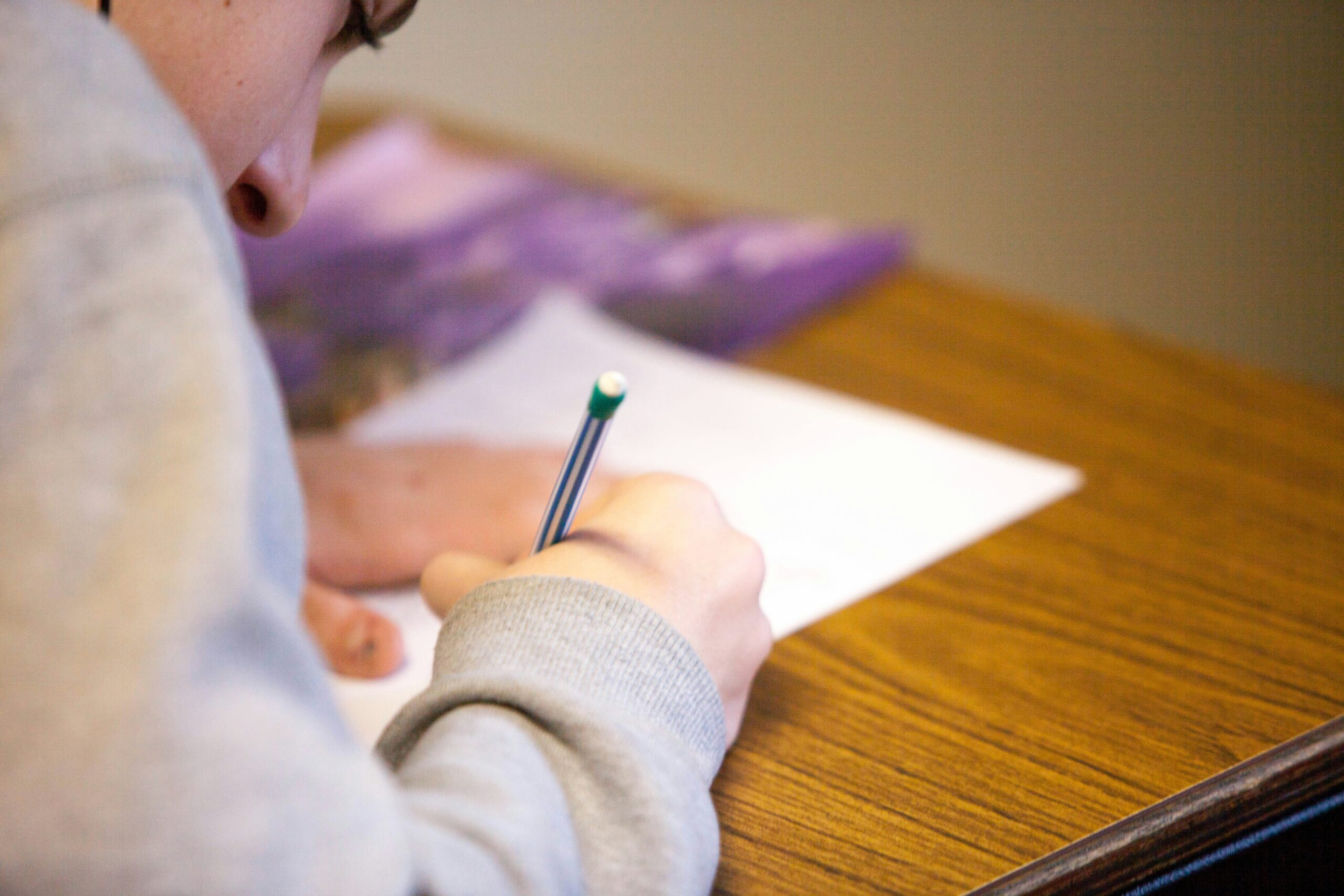 A person in a grey sweatshirt writes with a pencil on paper at a wooden desk.