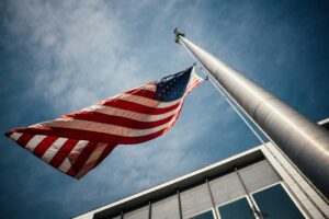 View from below of a U.S. flag waving on a tall flagpole against a blue sky, with a building partially visible.