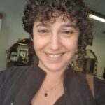 A person with curly hair smiles at the camera in a room with mirrors and salon equipment.