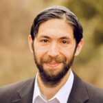 Man with a beard, wearing a suit and a kippah, smiles at the camera with a blurred nature background.