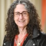 Person with curly hair and glasses smiling, wearing a leather jacket and necklace, in front of a neutral background.