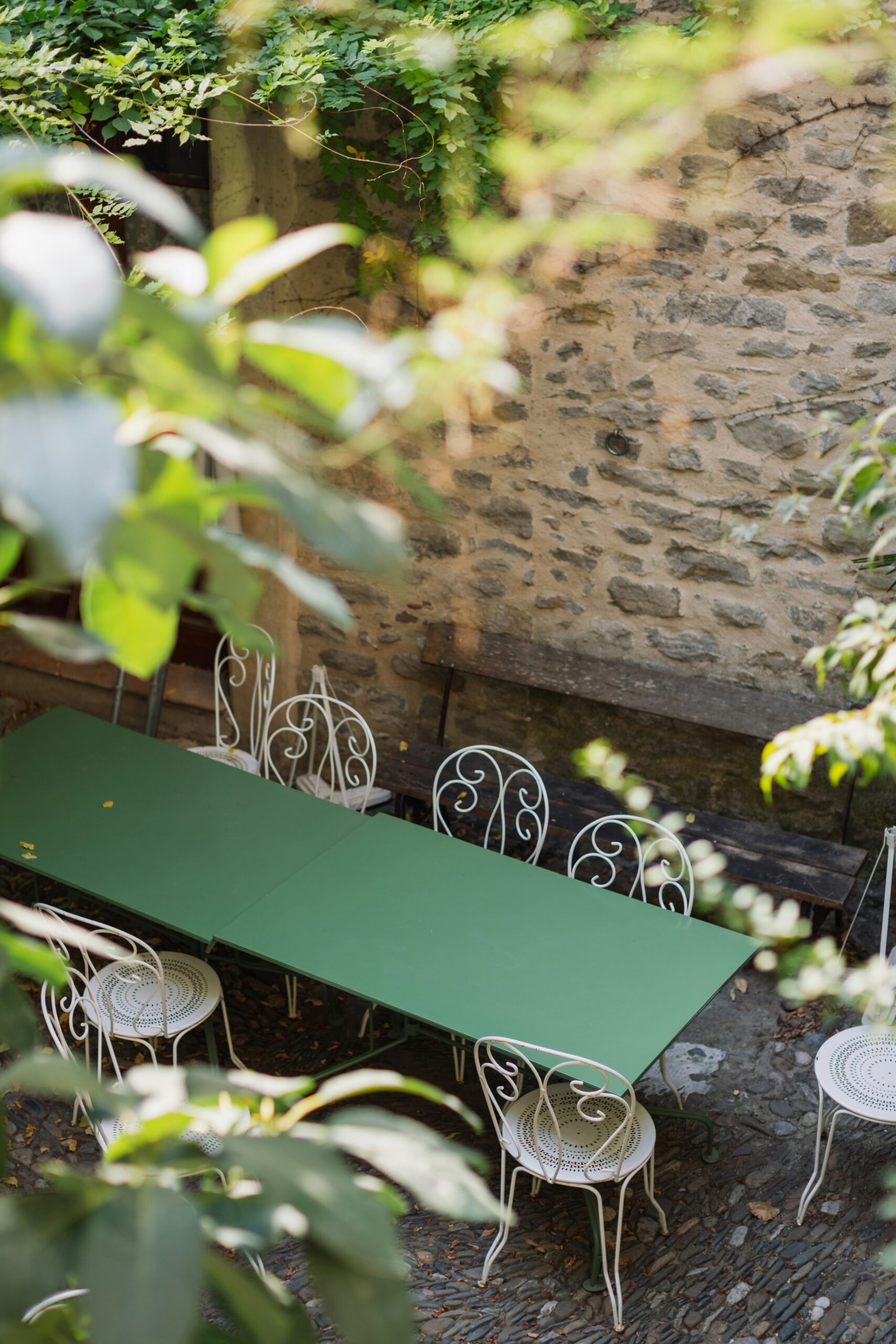 A green table with white chairs in a cozy outdoor courtyard surrounded by greenery and stone walls.