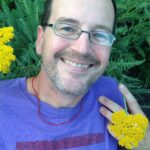 A man in glasses smiles while holding a yellow flower amidst green leaves.