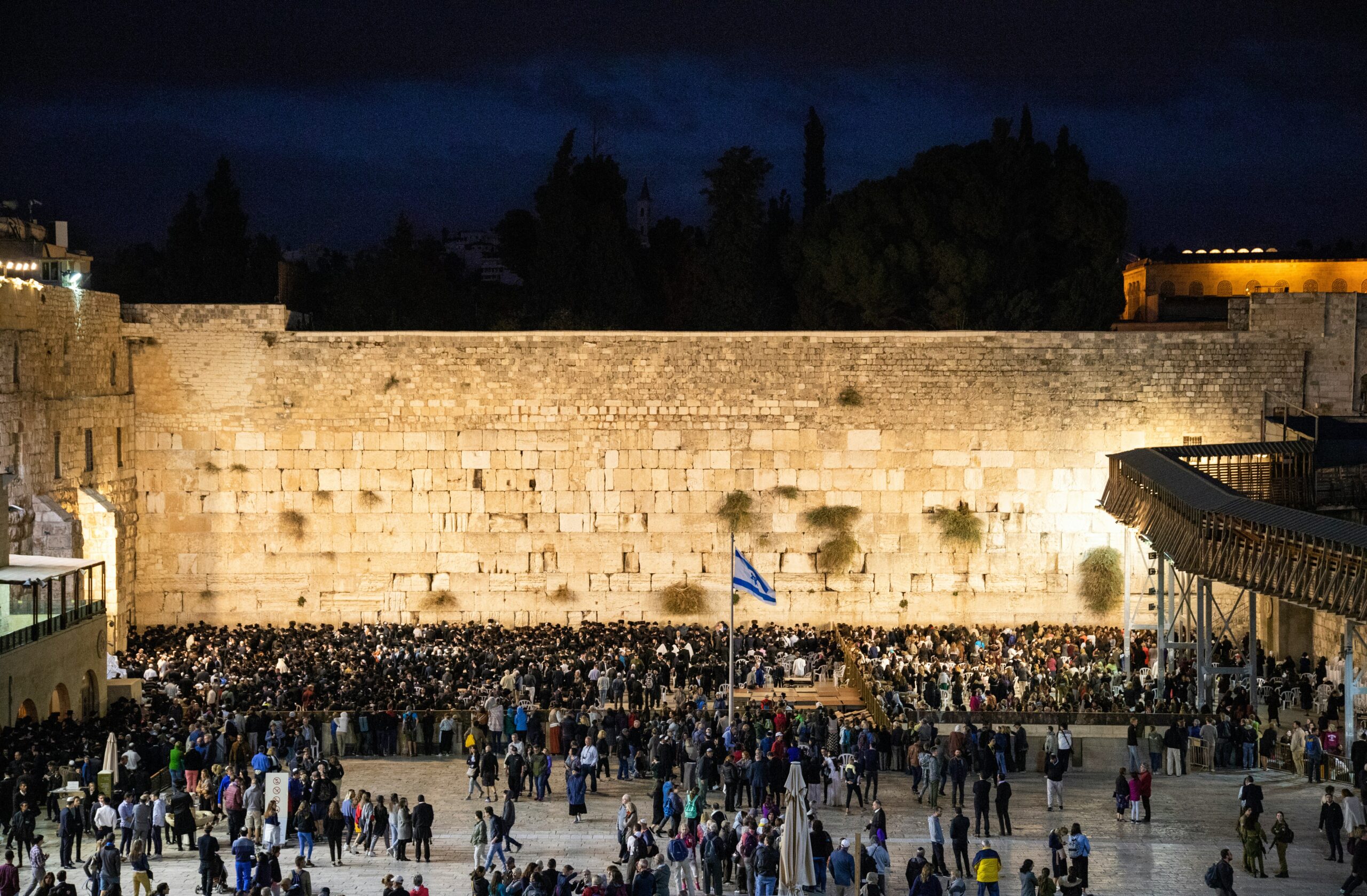A large crowd gathers for prayer at the Western Wall at night, with a dark sky above and a lit wall in the background.