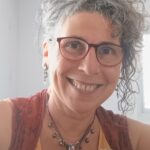 A woman with curly gray hair, wearing glasses, smiles at the camera in a bright room.