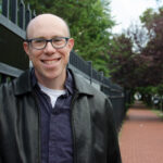 A smiling man in glasses and a leather jacket stands next to a wrought iron fence on a tree-lined sidewalk.