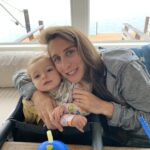 A woman hugging a baby in a high chair by a window overlooking a lake.