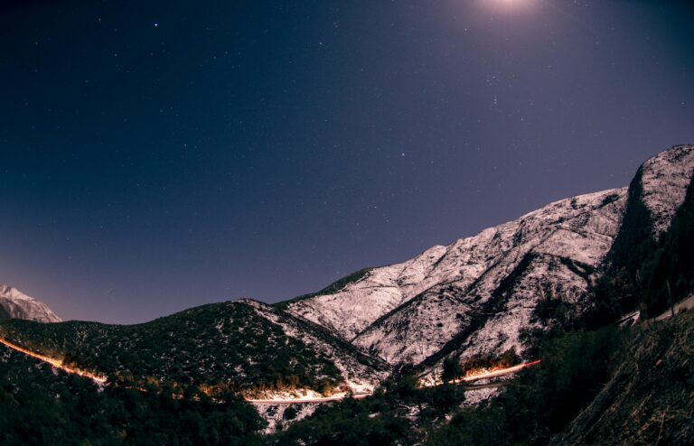 Starry night sky over snow-covered mountains with a faint light trail from a distant road below.