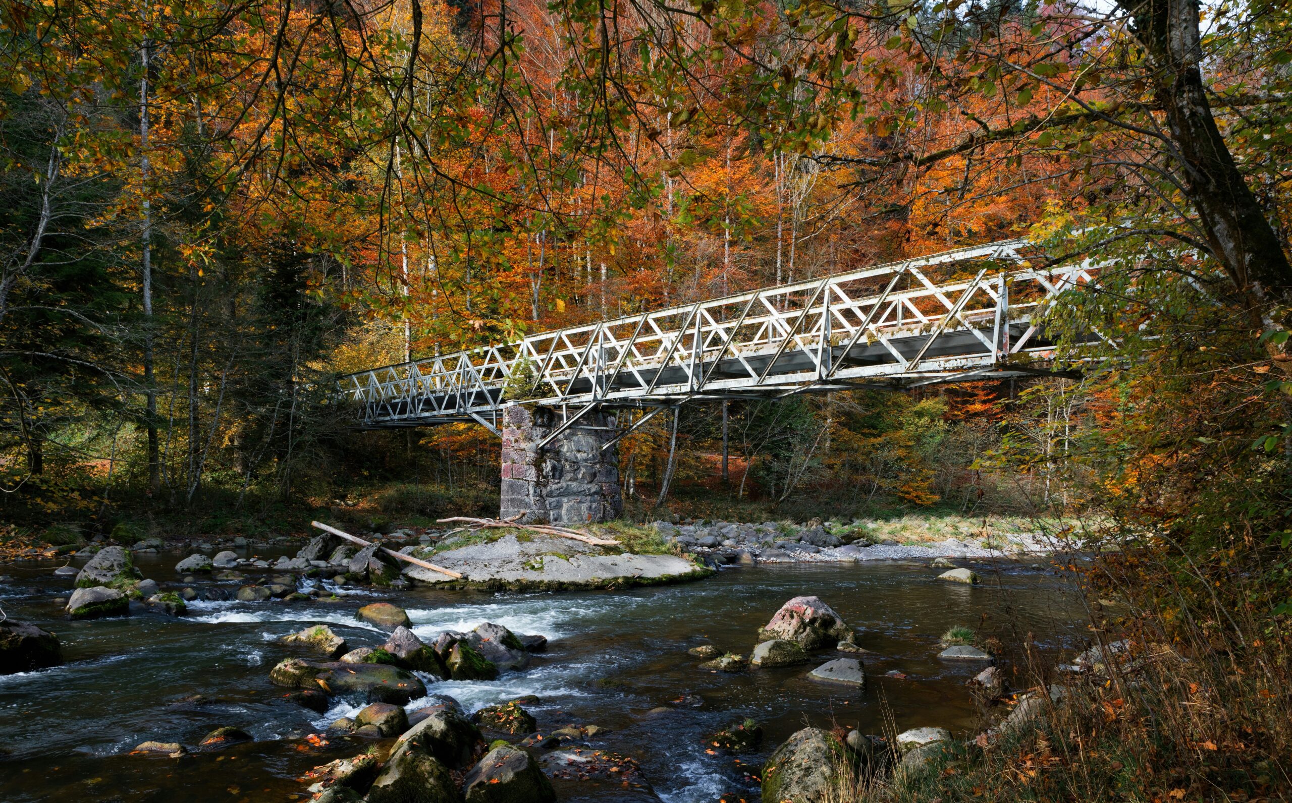 A metal bridge spans a rocky river in a forest with vibrant autumn foliage.