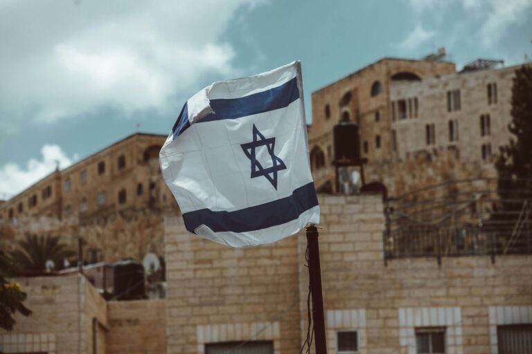 Israeli flag waving in front of historic stone buildings under a cloudy sky.