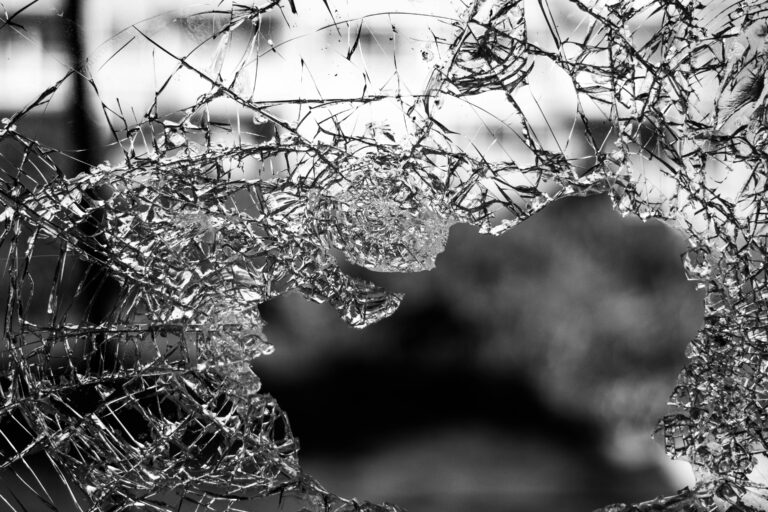 Close-up of shattered glass with jagged edges and a large hole, set against a blurred outdoor background.