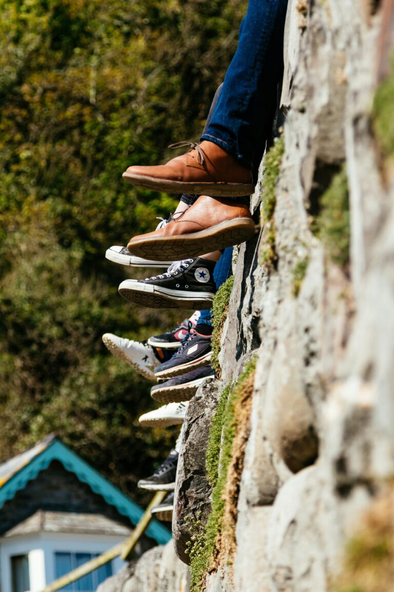 Six pairs of legs in different shoes dangle over a stone wall, with greenery and a house roof in the background.