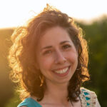 Smiling woman with curly hair, wearing a blue top, stands outdoors at sunset.