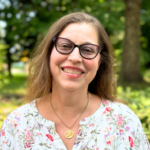 Smiling woman with glasses and floral top stands outdoors with trees in the background.