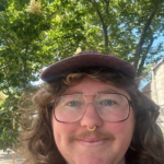 Person with long hair, glasses, and a cap smiles outdoors with trees in the background.