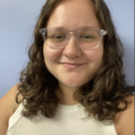 Smiling person with curly hair and glasses in a white top against a light blue wall.