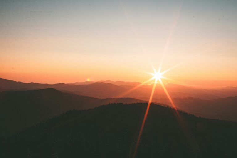 Sunrise over mountains with a bright sunburst, soft orange sky, and layered silhouettes of hills.