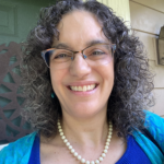 Woman with curly hair and glasses, smiling, wearing a blue top and pearl necklace, standing in front of a house.