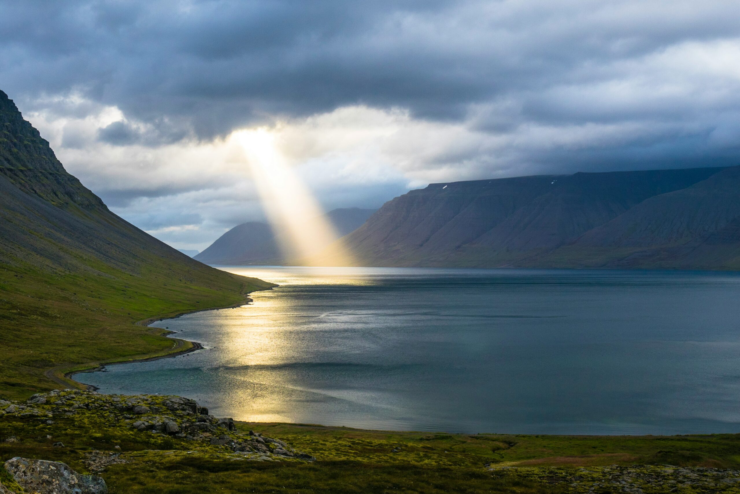 Sunbeam piercing through clouds over a serene fjord with mountains, creating a dramatic and peaceful scene.