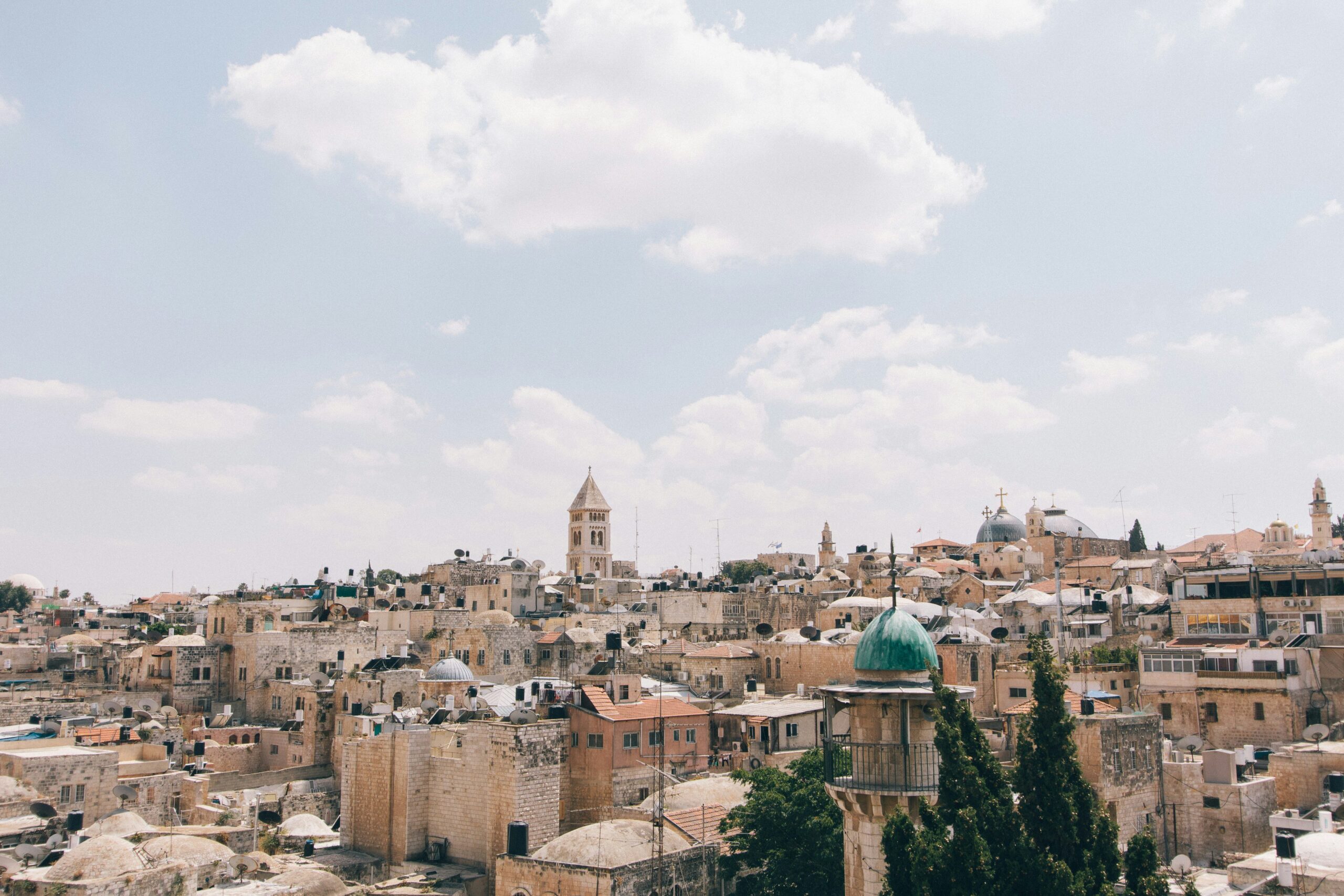 Skyline of an ancient city with beige stone buildings and a prominent church tower under a cloudy sky.