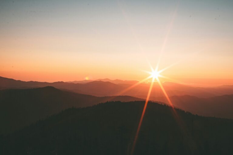 Sunrise over mountain ridges with a bright sunburst and warm orange and yellow hues in the sky.