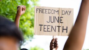 A hand holds a cardboard sign reading FREEDOM DAY JUNE TEENTH with raised fists nearby.
