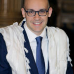 A smiling man in glasses, suit, and tie wears a white prayer shawl over his shoulders indoors.