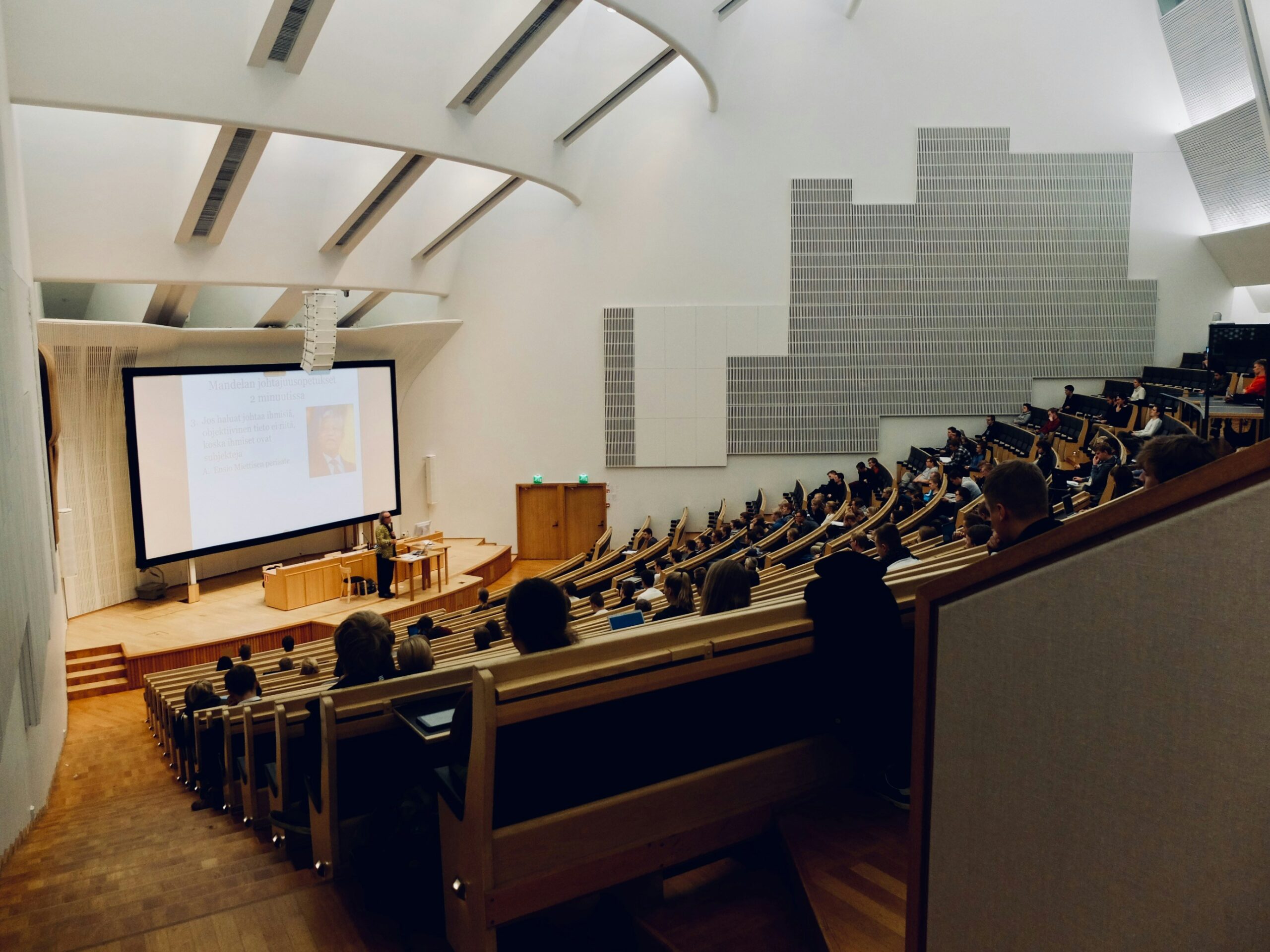 A large lecture hall with students seated and a presentation projected at the front.