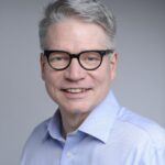 Smiling man with gray hair, glasses, and a light blue shirt against a plain gray background.