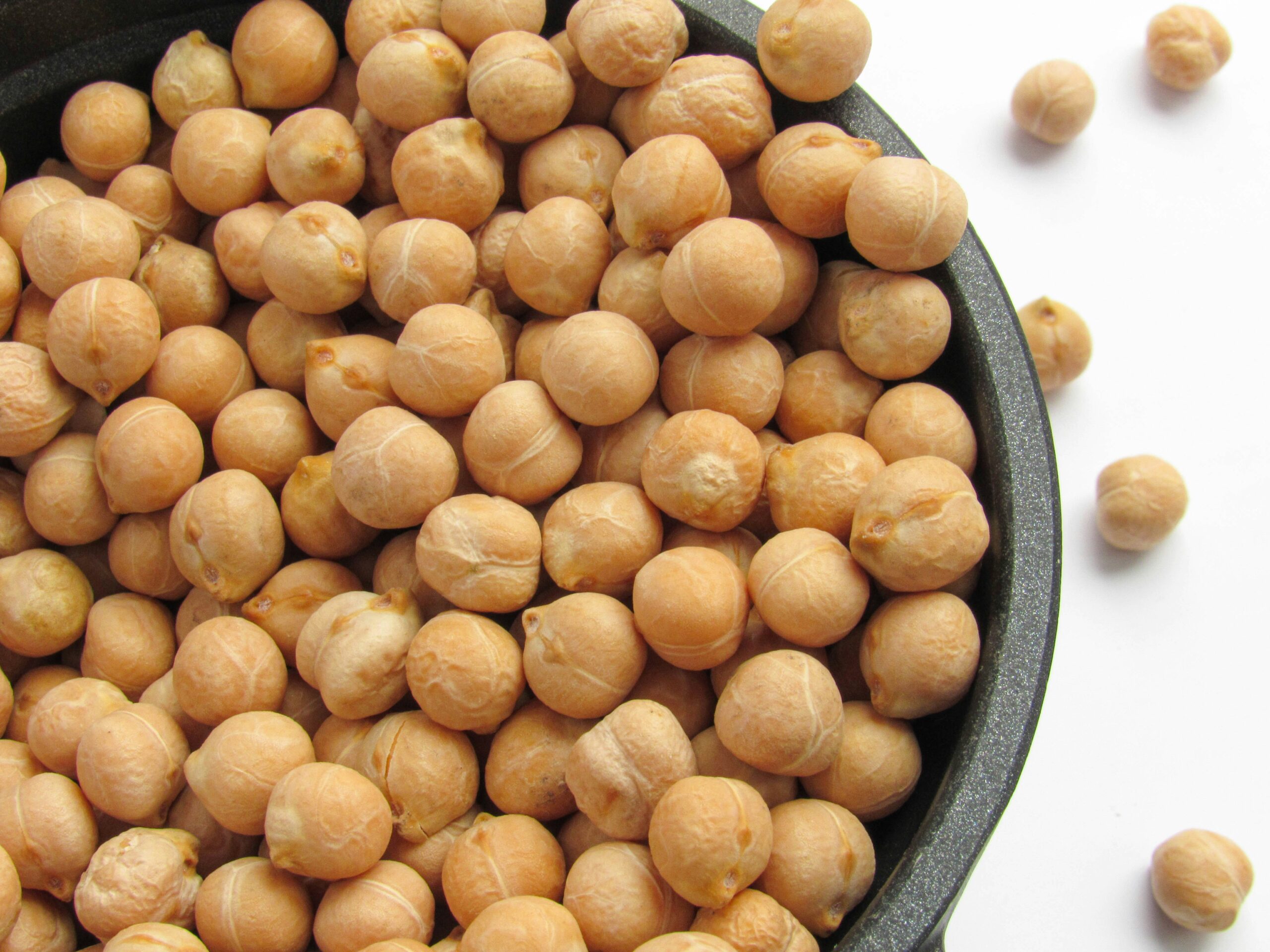 A black bowl filled with dry chickpeas, with a few chickpeas scattered on a white surface.